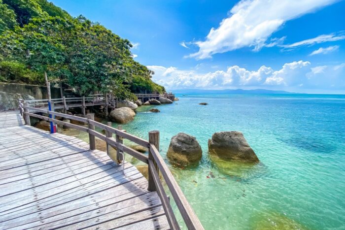 Wooden boardwalk along Koh Phangan coastline with turquoise sea, rocks and tropical greenery under blue sky
